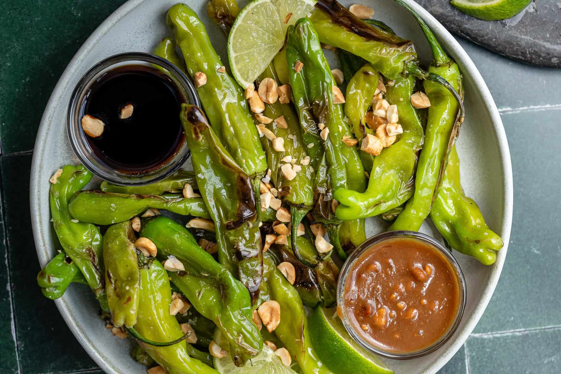 a large plate full of blistered shishito peppers with lime wedges, peanuts, and two bowls with sauce