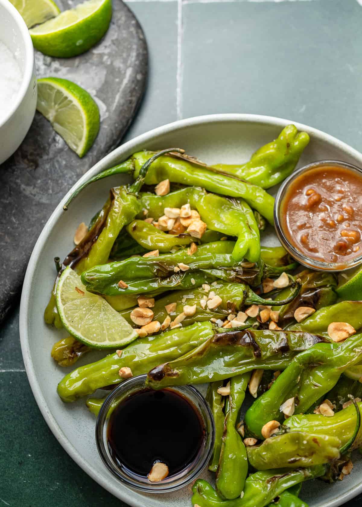 overhead shot showing blistered shishito peppers topped with peanuts, flaky salt, and lime wedges with sides of sauce